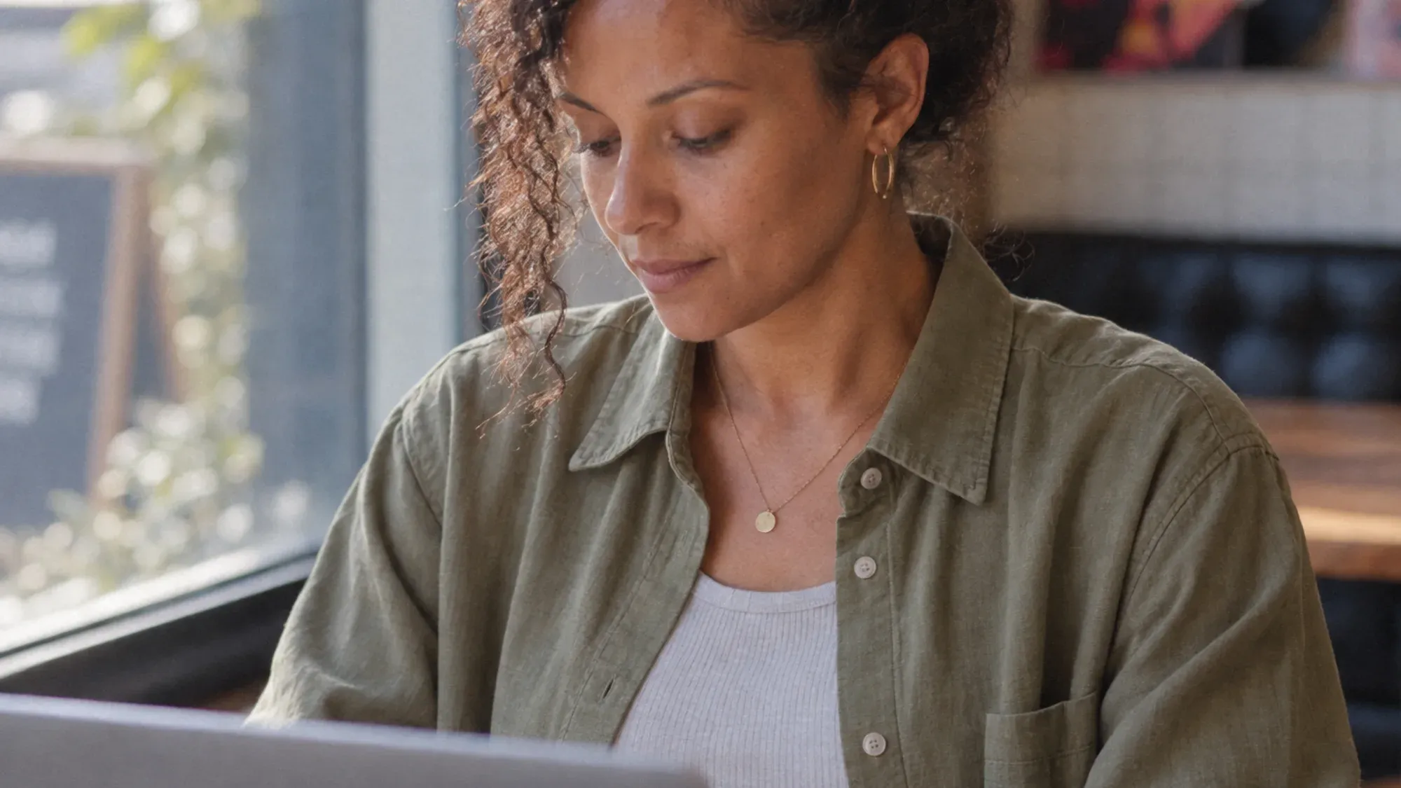 The woman in an olive shirt working on a laptop in a cafe, a close angle from the board.