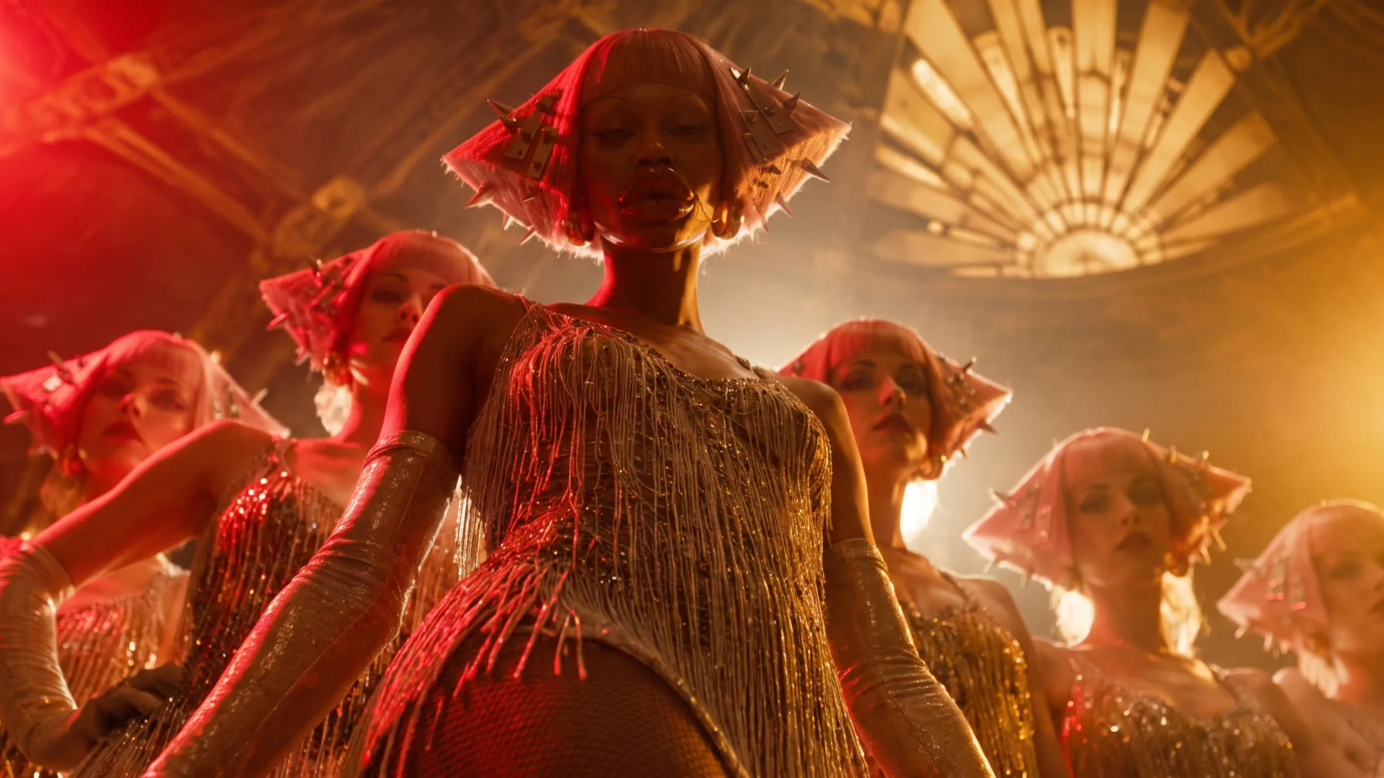 The same stage relit, red light falling from the upper left across the lead dancer and the line, gold backlight and haze behind them on the right.