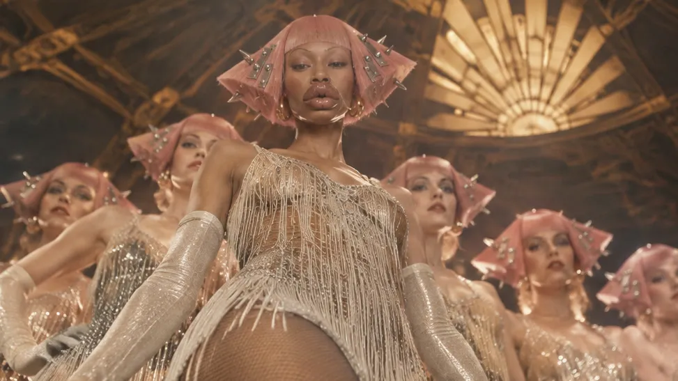 A line of dancers in pink spiked wigs and fringed silver costumes under a gilded Art Deco dome, the lead dancer in front, in soft warm stage light.