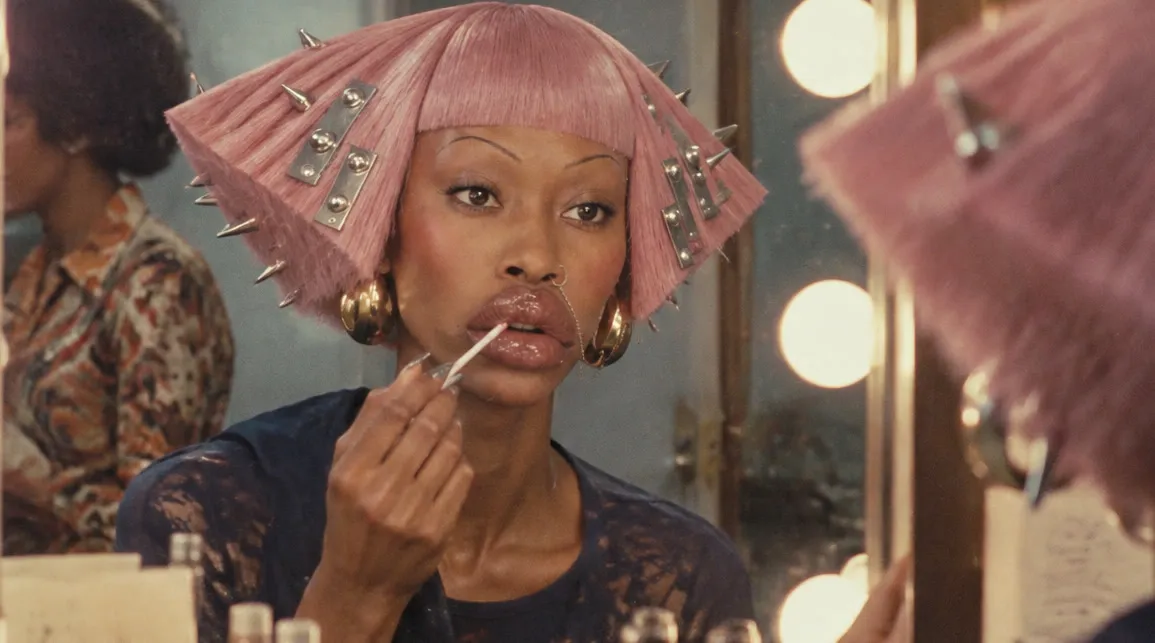 The character at a dressing room mirror in 1970, pink spiked wig and gold earrings, applying lip gloss under round bulb lights, in even light.
